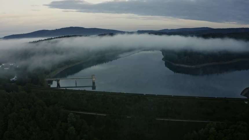 Structure links forested shores over lake surrounded by mist and gentle hills