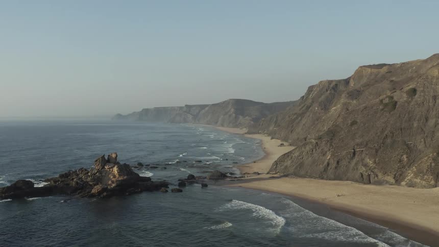 An aerial tracking shot moves along a rugged curving sandy beach backed by high dark cliffs where waves break around a dramatic rocky sea stack