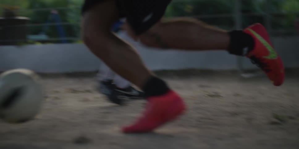 A person kicks a soccer ball on an outdoor dirt court while other players move in the background