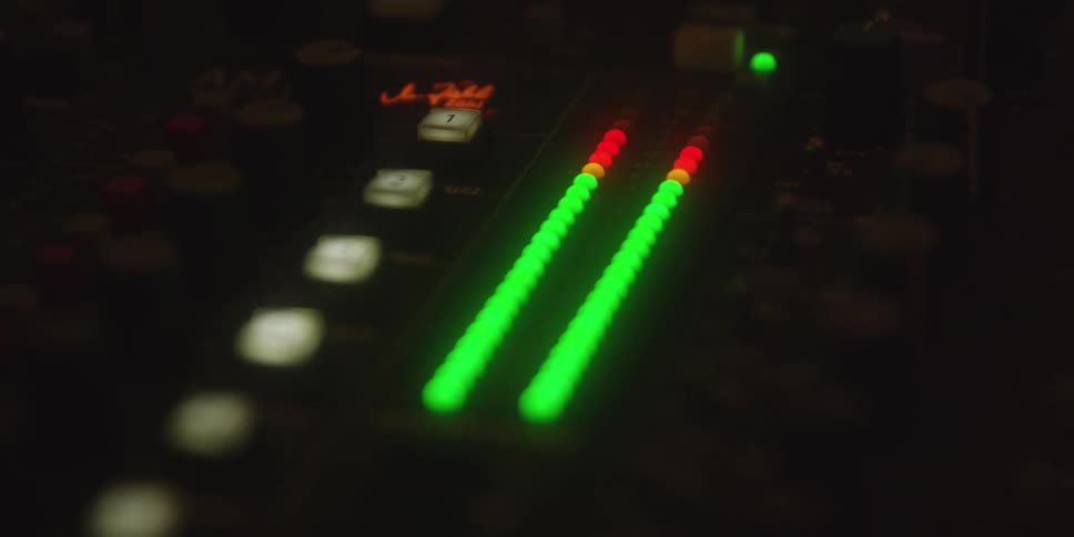 A closeup shot of a dark audio mixing console shows the bright green and red led level meters actively monitoring an audio signal which peaks and then drops off