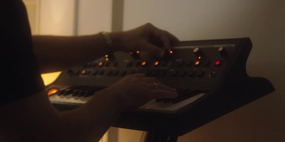 A person in a low light environment plays a black synthesizer keyboard with one hand while the other hand adjusts the control panel knobs highlighting the movement of the hands and the machines red indicator lights