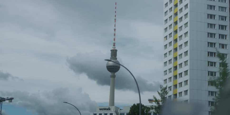 A giant tower stands prominently against a cloudy grey sky flanked by a tall modern apartment building and streetlights in the foreground