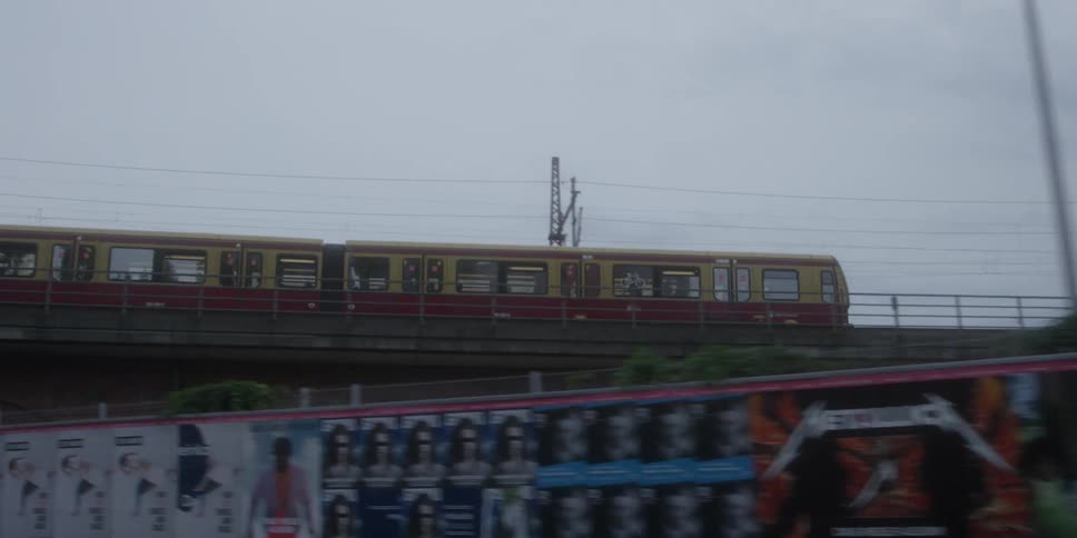 An elevated yellow and red tain passes lefttoright on an overpass under a gray sky and a parked truck on the street level below