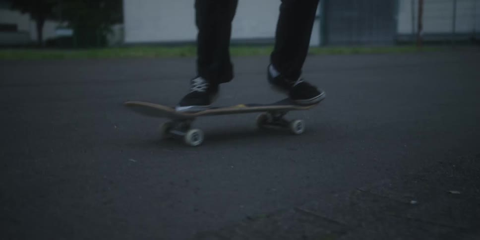 A person wearing black pants and sneakers performs a skateboarding ollie on a dark asphalt surface landing successfully before skating away from the camera
