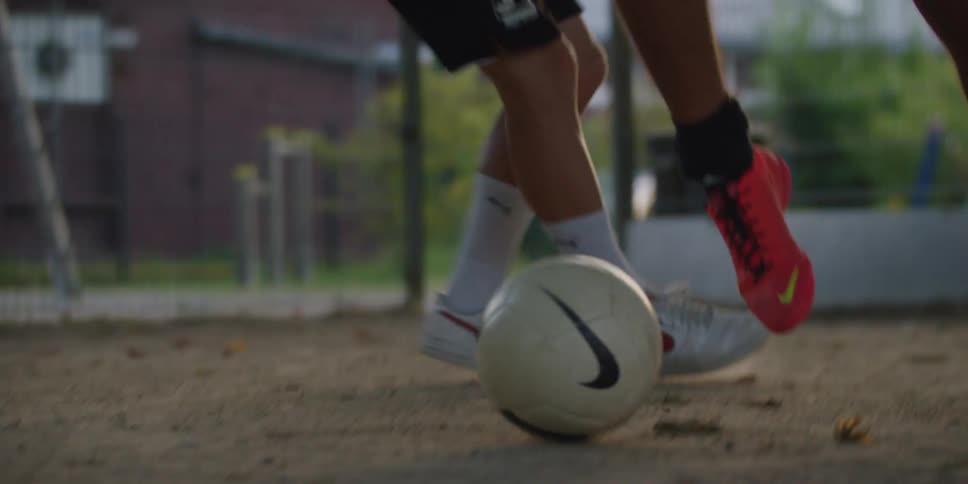 On a dusty court a players feet in bright cleats skillfully dribble a soccer ball evading another player who tries to defend