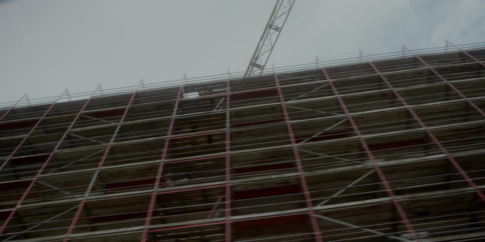 Structure under construction with red scaffolding and elevated crane beneath cloudy sky