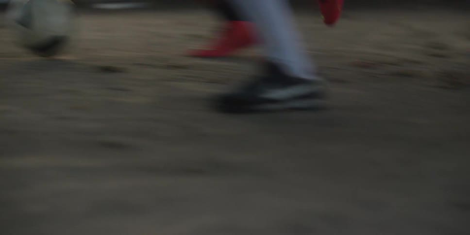 Players contest ball possession on dirt field with colorful cleats and dry leaves scattered around