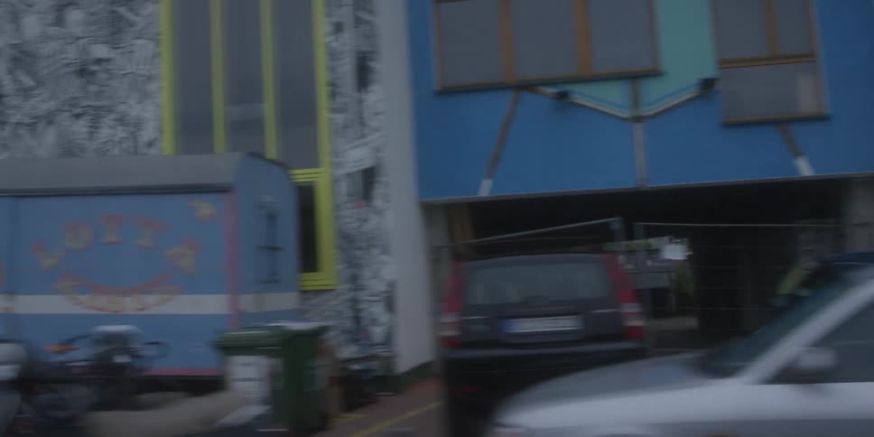 A colorful muralcovered building with a large stylized eye painting followed by a transition to a plain blue building as a car drives pasta mostly blackandwhite mural or graffiti wall with cars parked nearby and people walking set against an in