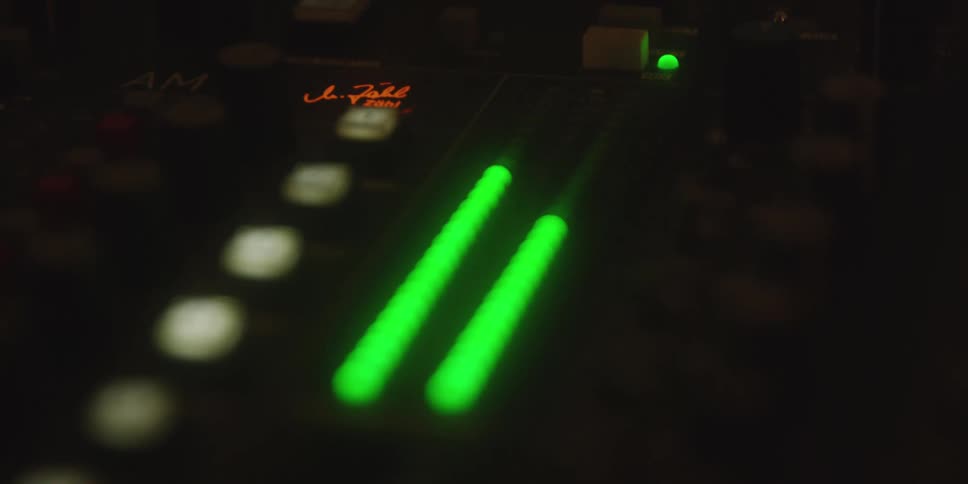 A dim studio soundboard glows with green and red level lights while the surrounding workspace sits in deep shadow