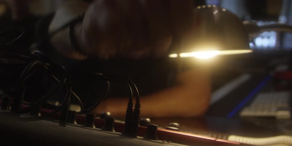 A man in a dim studio leans in and unplugs cords from audio equipment with a neutral focus while another person sits blurred in the background