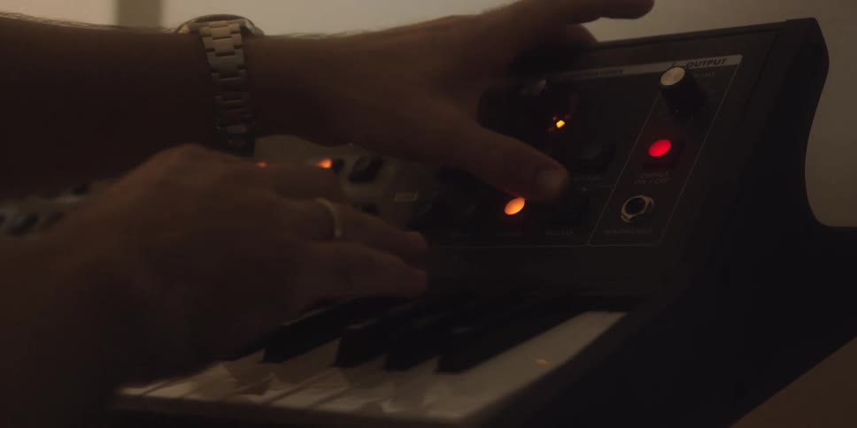 A musicians hands adjust the synthesizers control knobs while simultaneously playing the keyboard finetuning the sound in a dimly lit studio setting