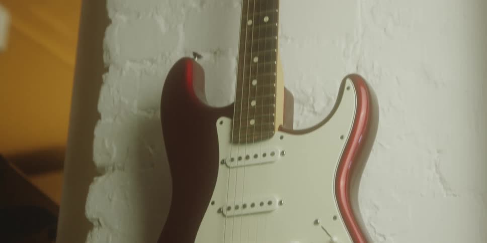 A maroon electric guitar is filmed in a closeup shot as it hangs vertically on a textured white wall in an indoor softly lit environment