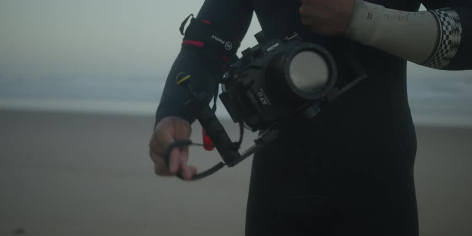A photographer in a wetsuit stands on the beach at dusk carefully securing a safety leash to their arm before gripping their large underwater camera rig