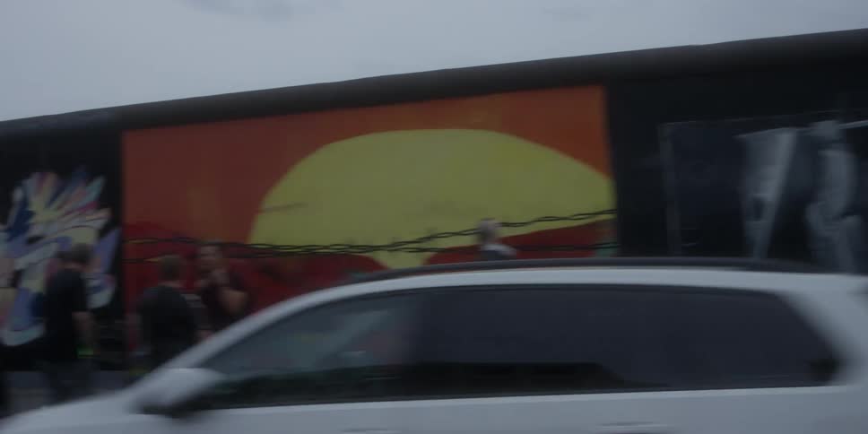 Moving alongside a long stretch of urban street the camera captures a historic concrete wall covered in diverse and colorful murals partially obscured by the flow of traffic passing in the foreground