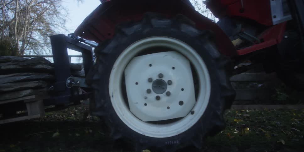 This is a lowangle closeup shot focusing on the large rear wheel of a small red tractor parked on a grassy patch of ground the tractor is hitched to a pallet loaded with stacked darkcolored flat stones or slate