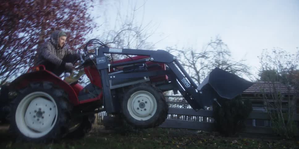 A man drives a tractor on a backyard lawn at midday while wearing a hoodie