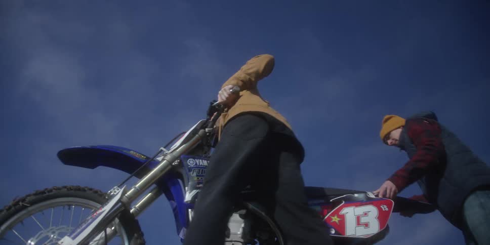 One person stands holding the handlebars of a motorcycle and the other person lifts the rear fender