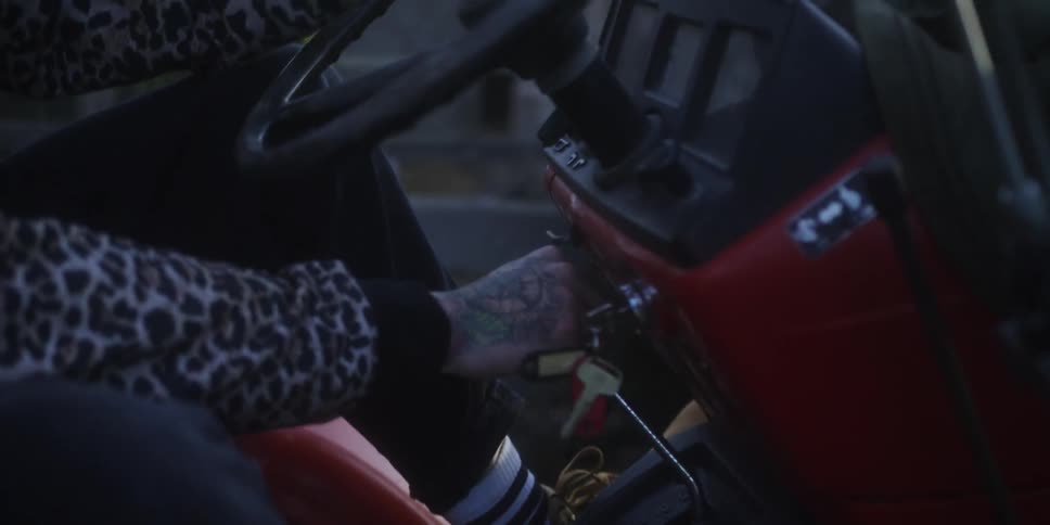 Tattooed hand turning ignition key on red tractor dashboard with visible steering wheel and switches