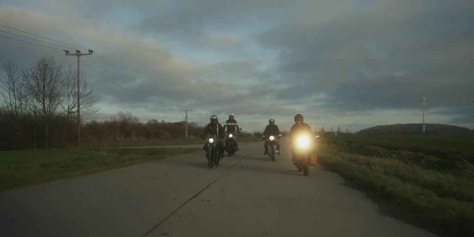 A group of four motorcyclists is riding down a paved country road at dusk the sky is cloudy and the bright headlights of the motorcycles illuminate the way as they pass fields and a power line