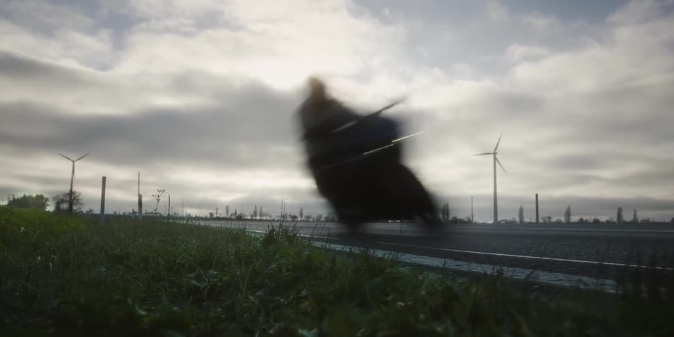 The video captured from a low angle near a roadside features a view of an empty twolane road stretching into the distance under a cloudy skyin the background there are several large wind turbines visible with their blades turning indicating a wi