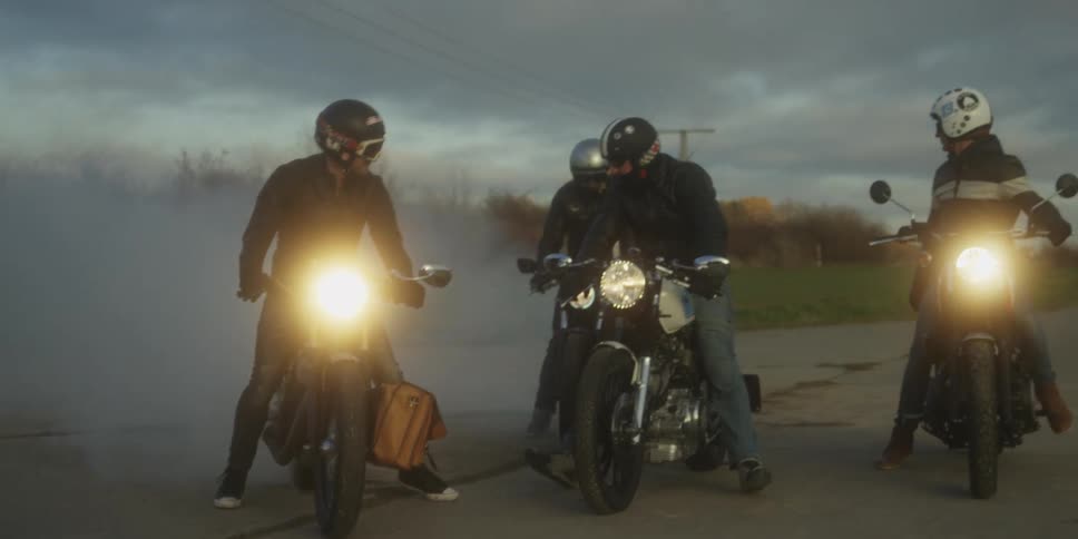 Four men on classic motorcycles sit revving their engines creating heavy tire smoke and illuminating the dark road with their bright headlights under a cloudy evening sky