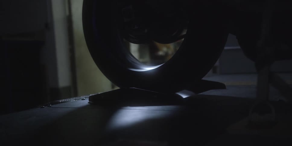In a dimly lit workshop a motorcycles rear wheel begins to spin rapidly on a dynamometer its motion highlighted by a single spotlight from below