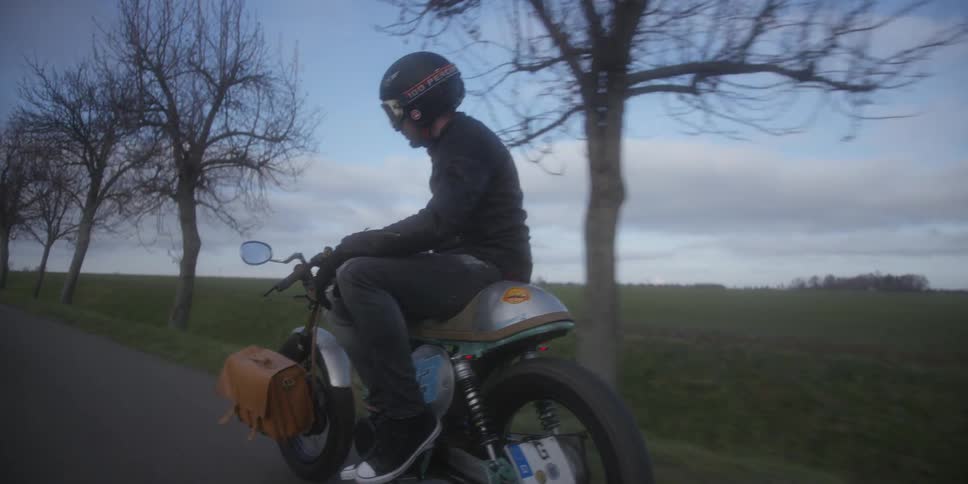 A motorcycle rider dressed in dark clothing helmet and goggles is sitting on a vintage or customized motorcycle with a small saddlebag attached looking directly at the camera while moving slowly down a paved country road