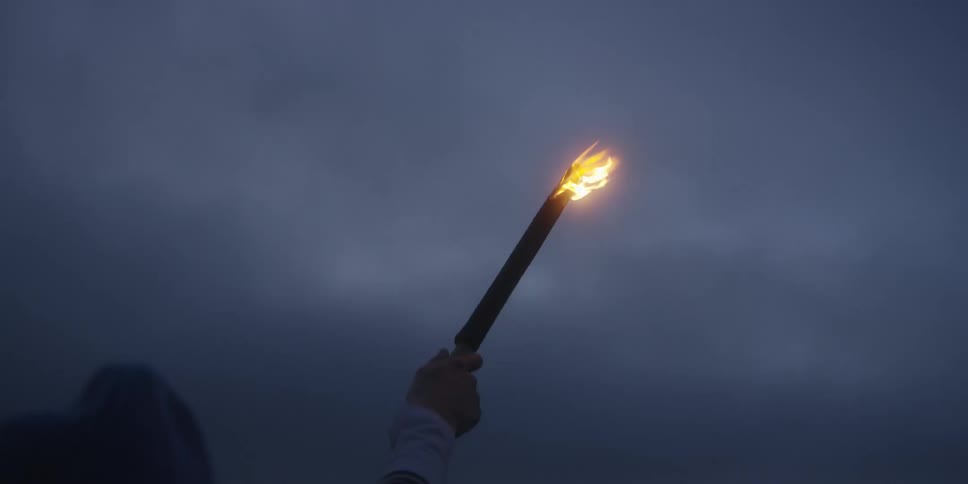 This is a shot from behind a person showing their outstretched arm holding a burning torch straight up against a uniformly dark overcast or twilight sky the flame is a small but clear burst of yelloworange the only bright element in the frame