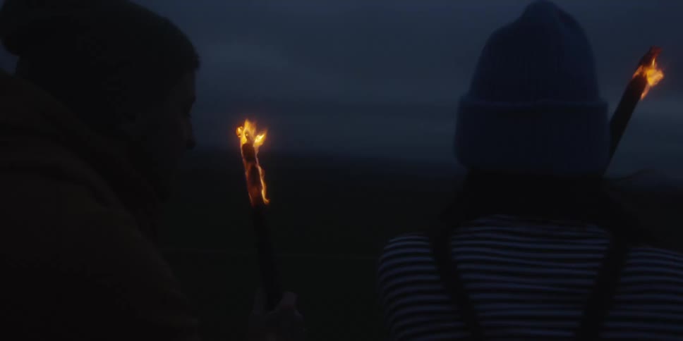 This video captures two people standing sidebyside in a dark outdoor environment possibly at dusk or night they are both holding up flaming torches which cast a warm flickering light on their faces and clothing