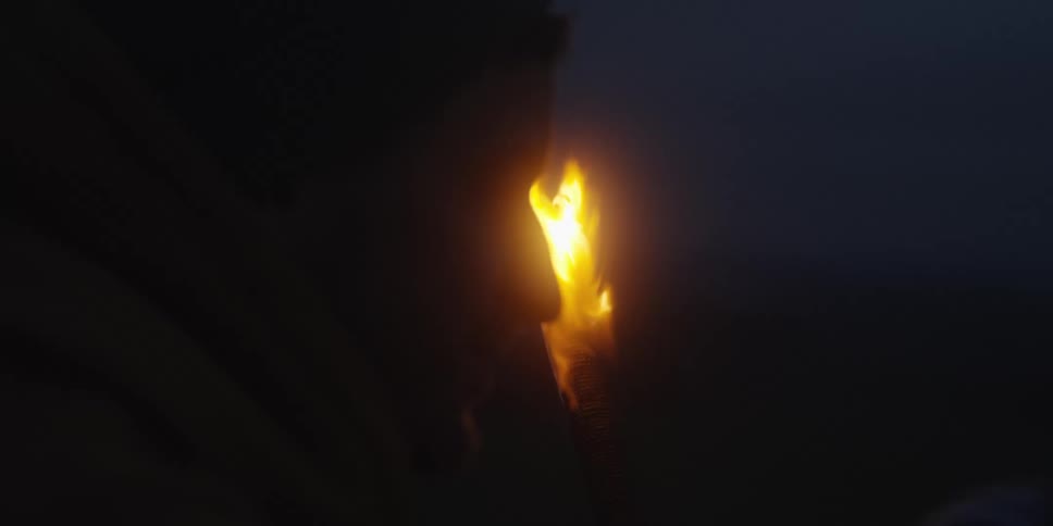 This video is a closeup profile shot of a person in a hooded garment viewed from the left side who is standing in a very dark setting the main source of light is a bright flickering torch flame which illuminates the persons face from the front righ