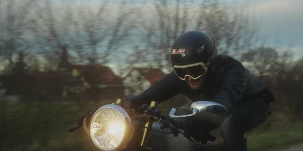 Two motorcyclists wearing black jackets and helmets with the lead rider wearing goggles are driving down a narrow road in a rural or suburban area possibly at dusk or in low light indicated by the motorcycles bright headlight