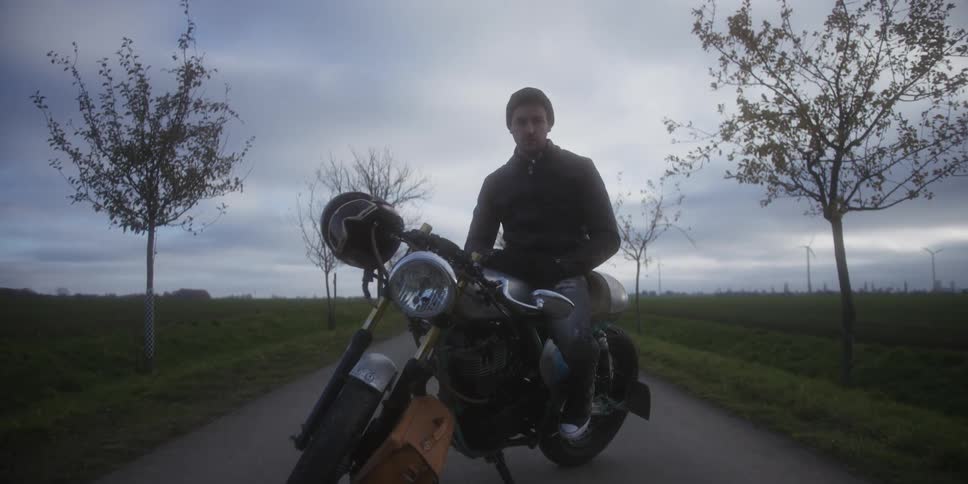 A man sits on a motorcycle in the middle of the road at midday wearing a beanie hat with a helmet resting on the handlebar
