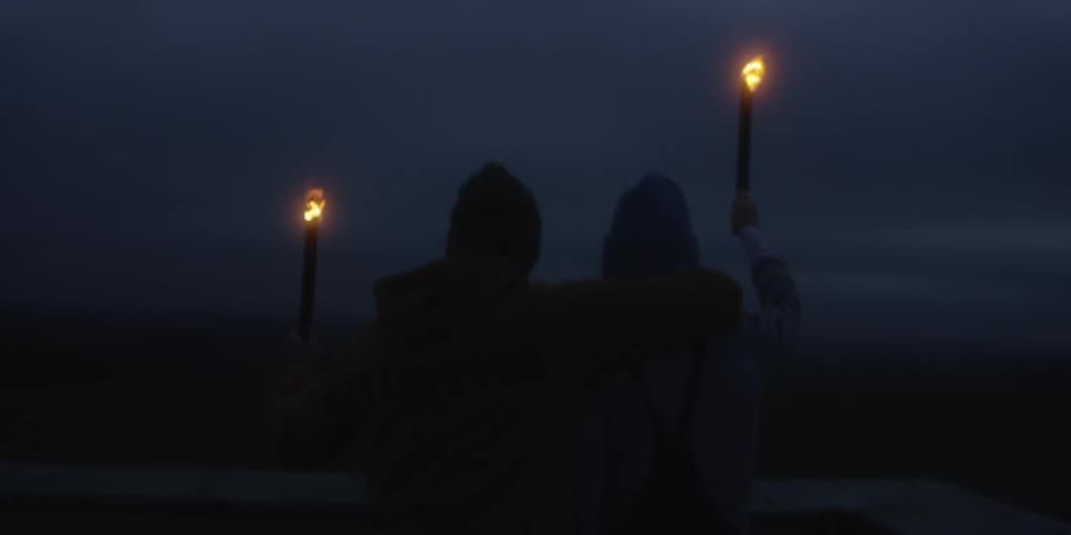 Two people on a balcony holding torches on their hands and lifting them up towards the sky outdoors during night time