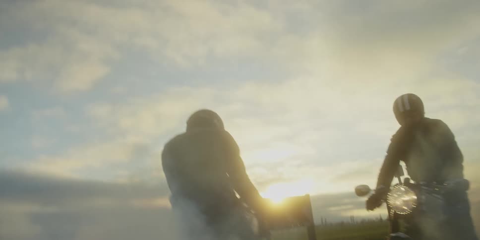 Two men ride a motorcycle at sunset revving the bike and creating a series of white smoke