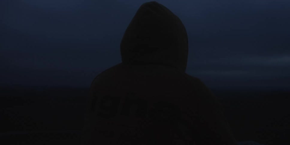 A person wearing a large yellow hooded jacket is seen in a dark silhouette against a cloudy blueblack sky turning their head slightly to look into the distance