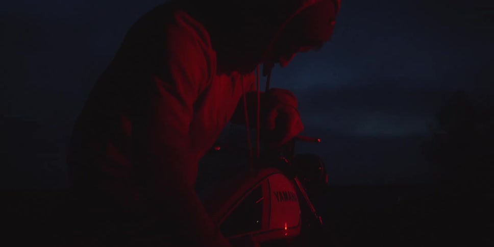 A person in a hooded sweatshirt sits on a yamaha motorcycle in the darkness illuminated solely by a red light that casts a deep silhouette as they lean down seemingly preparing to start the bike at night
