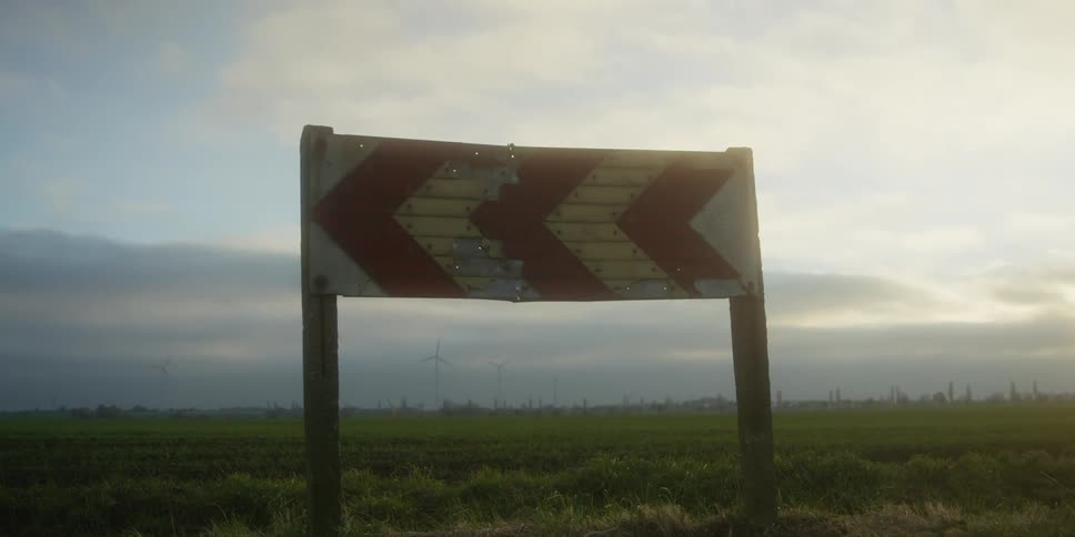 Sign on the side of a rural road with wind turbines and buildings in the distance