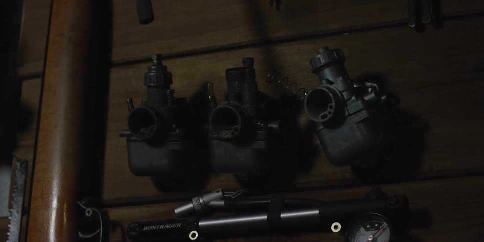 Three carburetor parts rest on a wooden workbench under dim light