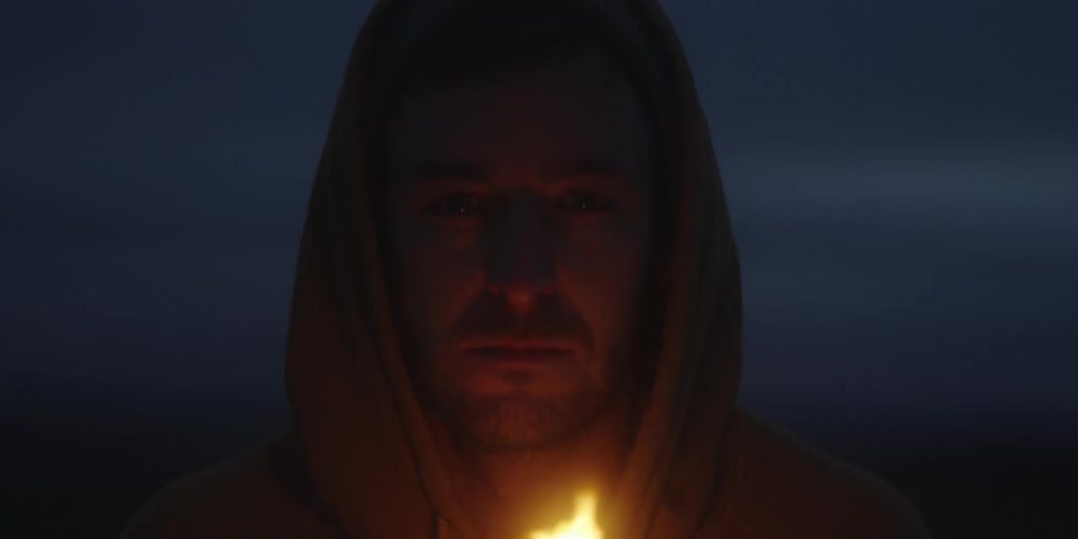 Hooded man lit by a flame close to his face in an outdoor nighttime setting