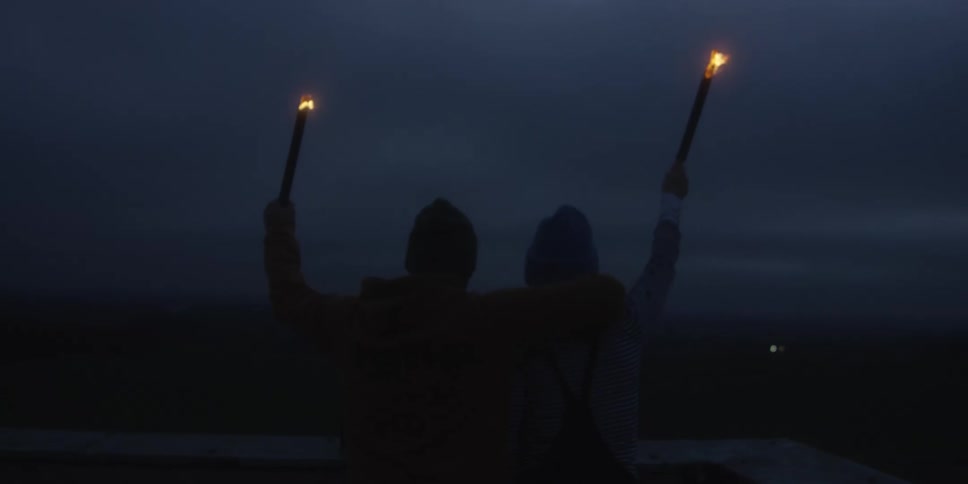Embracing with their backs to the camera two people triumphantly hold flaming torches high against the deep twilight sky