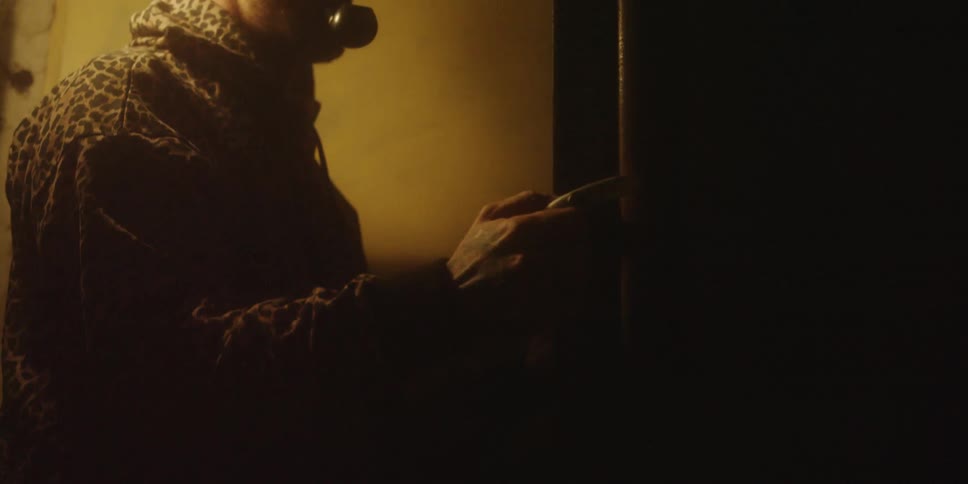 A man wearing a beanie and a leopard print jacket walks into a dim confined room with a large wooden pipe in the moody yellow light