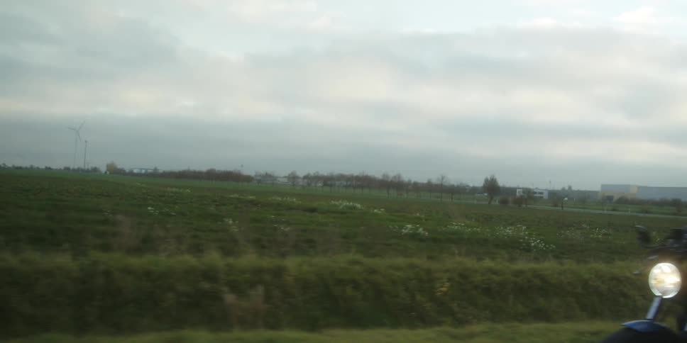 Two people wearing a leather jacket and jeans rides a custom motorcycle through a green rural field on a cloudy day