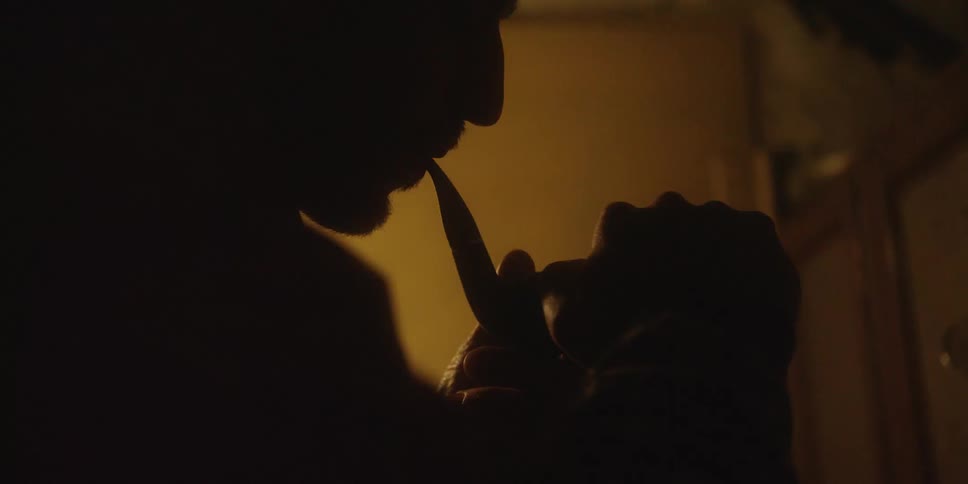 The video shows a man in a dimly lit dark setting attempting to light a tobacco pipe he is seen in closeup profile silhouetted against a faint background light with a distinct glow coming from the small flame of his lighter as he brings it near the b