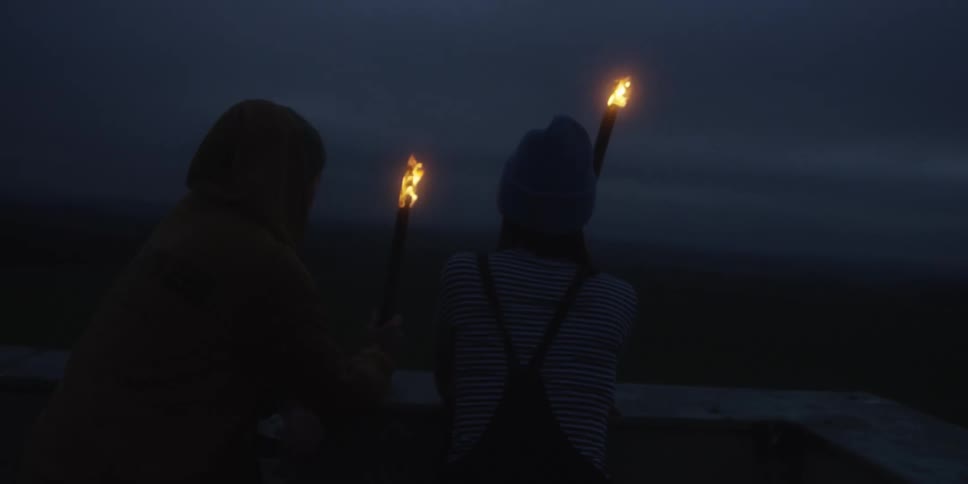 Two people one in a yellow hood and the other in a blue beanie sitting on a platform holding flickering torches against a dark featureless night sky