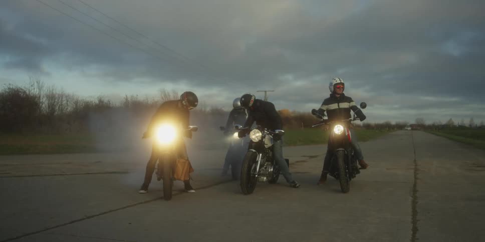 A group of four motorcycle riders wearing helmets and gear are gathered on a quiet rural road under an overcast sky their bikes headlights are illuminated and a slight haze or smoke is visible around the riders and their motorcycles
