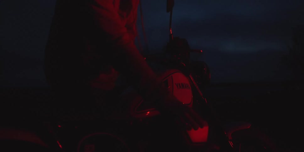 A man wearing a hoodie is out in the woods at night with his motorcycle as a campfire illuminates the surroundings