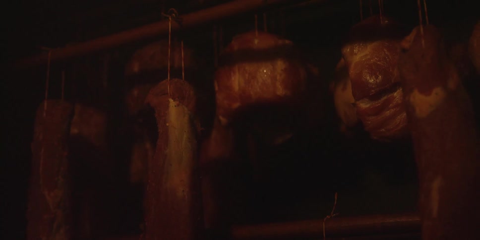 Several large pieces of meat likely pork hang from a wooden rack in the dark smoky interior of a smokehouse during the curing and preservation process
