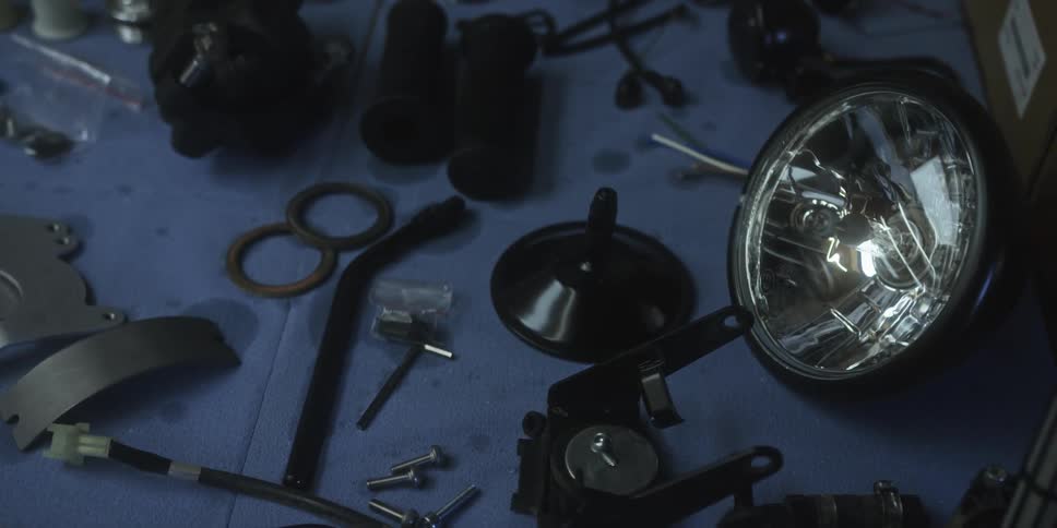 The video shows a closeup dimly lit shot of various motorcycle parts scattered on a blue surface likely a workbenchcomponents visible include a headlight assembly black grips several metal brackets and bolts wiring and a small wrench