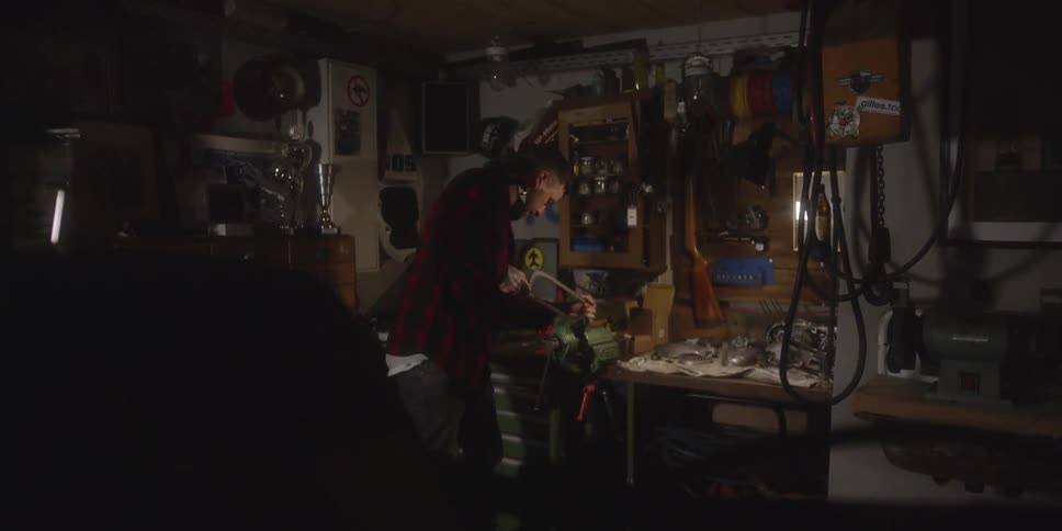 A man works with tools at a cluttered workbench inside a dimly lit garage