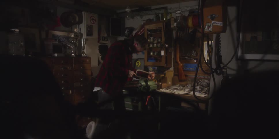 A man wearing a red plaid shirt uses a hacksaw to cut a piece of metal held in a bench vice in a crowded dimly lit workshop filled with tools and miscellaneous items